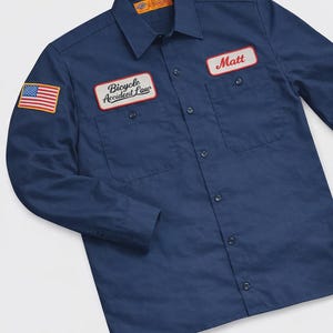Könnte beinhalten: Marineblaues Arbeitshemd mit einem amerikanischen Flaggenaufnäher und zwei rechteckigen Aufnähern. Ein Aufnäher lautet "Bicycle Accident Law" und der andere "Matt". Das Hemd hat zwei Brusttaschen und Knopfverschlüsse.