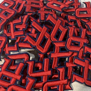 Könnte beinhalten: Ein Haufen roter, gestickter Buchstabenaufnäher mit marineblauem Rand. Die Aufnäher sind in einer stilisierten Schriftart gehalten, wahrscheinlich für die Individualisierung von Kleidung oder Bastelprojekten. Das Bild zeigt eine Nahaufnahme der Aufnäher.