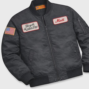 Op de afbeelding: Zwarte bomberjack met ritssluiting, geribde manchetten en kraag. De jas heeft een Amerikaanse vlag patch, een patch met de tekst "Bicycle Accident Law", en een patch met de tekst "Matt".