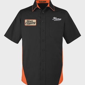 Può includere: Camicia da lavoro nera a maniche corte con dettagli arancioni sui lati, sul colletto e sui bordi. Presenta una toppa con la scritta "Your Company" e la parola "Name" in corsivo bianco. Un classico stile uniforme.