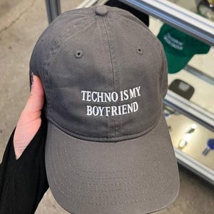 May include: A gray baseball cap with the text "TECHNO IS MY BOYFRIEND" in white block letters. The cap has a curved brim and a fabric strap closure. The hat is made of a soft, durable material.