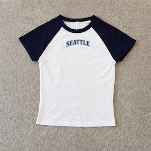 May include: White and navy blue raglan-style t-shirt with the word "SEATTLE" in light blue lettering across the chest. The shirt has a crew neck and short sleeves. The shirt is made of a soft, comfortable material.