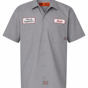May include: A gray short-sleeved work shirt with two chest pockets and a button-down front. The shirt has two patches: one that says "Bicycle Accident Law" and another that says "Matt". The Dickies logo is visible.