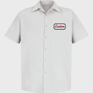 Op de afbeelding: Een lichtgrijs overhemd met korte mouwen, een kraag en knoopsluiting. Het heeft een borstzak met het woord "Custom" in rood schrift in een zwart-witte rand. Het overhemd heeft een klassieke werkkledingstijl.