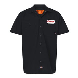 Peut inclure: Chemise de travail noire à manches courtes avec deux poches de poitrine et une fermeture boutonnée. Un écusson blanc avec le mot "Custom" en rouge est sur la poitrine droite. La chemise a un col classique et un petit logo en bas.