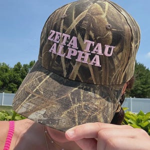 May include: A camouflage baseball cap with the embroidered text "ZETA TAU ALPHA" in pink. The hat features a curved brim and a classic six-panel design, suitable for outdoor activities.