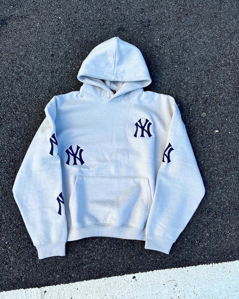 Puede incluir: Una sudadera con capucha gris claro con un bolsillo delantero y una capucha. El logotipo de los Yankees de Nueva York en azul marino est&aacute; presente en el pecho y las mangas. La sudadera est&aacute; hecha de un material suave.