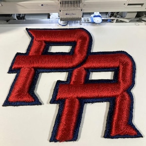 Può includere: Toppe ricamate con lettere rosse e blu navy, forse per una squadra sportiva o abbigliamento. Le lettere "PR" sono cucite con un aspetto strutturato, mostrando i dettagli del ricamo. Su una superficie bianca.