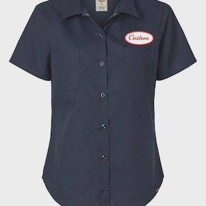 Op de afbeelding: Marineblauwe korte mouwen werkshirt met twee borstzakken en een knoopsluiting. Een ovale patch met het woord "Custom" in witte letters en een rode rand staat op de linkerborst. Het shirt heeft een klassieke kraag en is gemaakt van een duurzame stof.