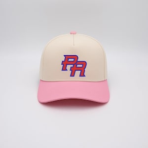 Puede incluir: Gorra de béisbol color crema con visera rosa. La gorra presenta las letras bordadas "PR" en azul con contorno rojo. La gorra tiene un botón rosa en la parte superior.
