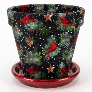 May include: A tapered flower pot with a black background featuring a pattern of red cardinals, green pine branches, pinecones, and gold stars, accented by white dots resembling snow. The pot sits on a matching glossy red circular saucer.