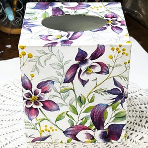 May include: A white tissue box featuring a floral design with purple, white, and yellow flowers and green leaves. The box has an oval opening on top for tissue dispensing. The floral pattern covers the entire box.