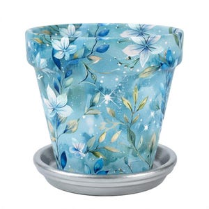 May include: A tapered ceramic flower pot featuring a watercolor-style pattern of light blue and white flowers with green leaves on a teal background, accented by small white sparkles. The pot sits on a solid, smooth, metallic silver saucer.