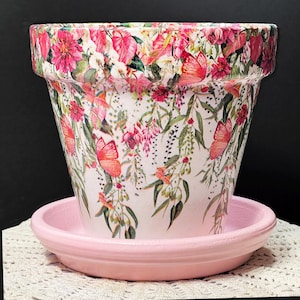 May include: A decorative flower pot with a matching saucer. The pot is white with a floral and butterfly pattern in pink, red, and green. The saucer is solid pink. The pot is approximately 7 inches in diameter and 6 inches tall.
