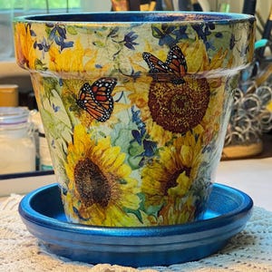 May include: A decorative plant pot and matching saucer featuring a vibrant sunflower and monarch butterfly pattern. The exterior displays yellow sunflowers with green foliage and orange butterflies on a light background, while the rim and saucer are painted in a solid metallic blue finish.