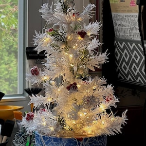 May include: A small, white Christmas tree with warm white lights, red berries, and silver ornaments. The tree is in a blue ceramic pot with a winter scene design. The tree is approximately 46 cm tall.