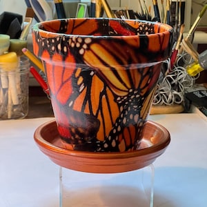 May include: A terracotta-colored flower pot and matching saucer, decorated with a vibrant monarch butterfly pattern in shades of orange, black, and white. The pot sits on a clear stand, showcasing the detailed butterfly design.