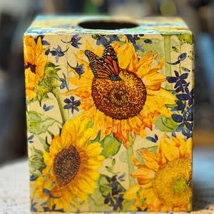 May include: A square tissue box with a floral design of sunflowers and a monarch butterfly. The box is decorated with yellow, orange, and green colours, creating a cheerful and decorative accent for any room.