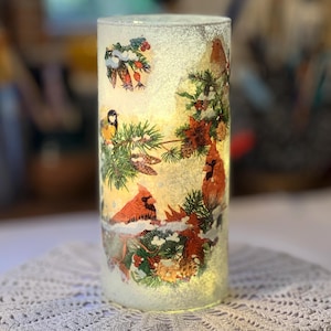 May include: A cylindrical glass candle holder with a winter scene featuring red cardinals, pine branches, and snow. The holder is illuminated from within, creating a warm glow. The design includes a frosted effect, enhancing the winter theme.