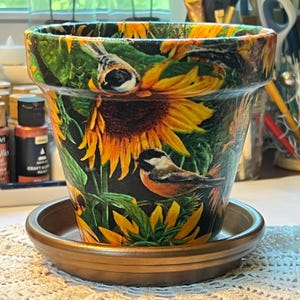 May include: A decorative terracotta flower pot with a matching saucer. The pot features a vibrant design of sunflowers and small birds. The colour palette includes yellow, green, brown, and black.