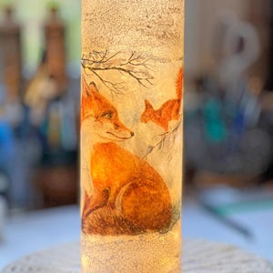 May include: A cylindrical decorative lamp featuring a winter scene with a fox and squirrel. The lamp has a textured, off-white surface with an illustration of the animals in shades of orange and brown. The lamp is lit from within, creating a warm glow.