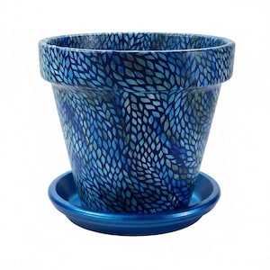May include: A blue ceramic flower pot with a matching saucer. The pot has a leaf-like pattern in shades of blue and green. The pot is designed for planting flowers or other plants.