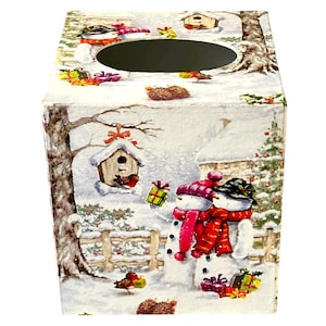 May include: A decorative tissue box featuring a winter scene with snowmen, birdhouses, and wrapped gifts. The box has a white background with illustrations of a snowy landscape, trees, and a wooden fence. The snowmen are wearing red scarves and hats.