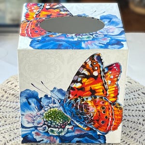 May include: A decorative tissue box with a butterfly design. The box features orange, red, and yellow butterflies set against blue flowers and a cream-coloured background. The box is a square shape.