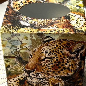 May include: A decorative tissue box featuring a detailed leopard print design. The box is rectangular with an oval opening on top. The leopard's face is prominently displayed, with brown and black spots.