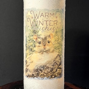 May include: A cylindrical candle holder with a frosted white exterior. The design features a winter scene with a small animal and the text "Warm Winter Wishes" in a decorative font. The candle holder is suitable for seasonal decor.