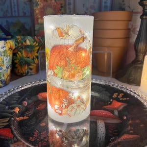 May include: A cylindrical, frosted glass candle holder with a fall harvest design. The design features a large orange pumpkin, smaller pumpkins, and a bird. The candle holder is illuminated with small, warm white lights, creating a cozy ambiance.