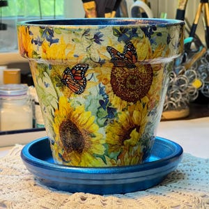 May include: A decorative flower pot with a matching saucer. The pot features a sunflower and butterfly design with yellow, orange, and blue hues. The saucer is a solid blue color. The pot is designed for planting flowers.