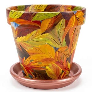 May include: A tapered terracotta plant pot featuring a hand-painted design of overlapping autumn leaves in vibrant shades of yellow, orange, red, and green. The pot sits on a matching metallic copper-colored circular saucer with a raised rim.