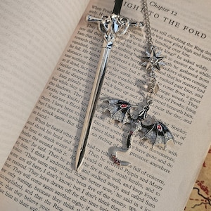 May include: A silver bookmark with a sword design and a chain, featuring a bat and star charm, lies on a book. The bookmark is detailed and elegant, ideal for fantasy readers.