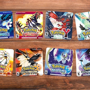 May include: Eight Pokemon video game cases are displayed on a wooden surface. The cases feature colorful artwork and the game titles "Alpha Sapphire", "Omega Ruby", "Y", "X", "Sun", "Ultra Sun", "Moon", and "Ultra Moon".