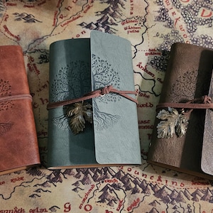 May include: Three leather journals in shades of brown and green, each with an embossed tree design. They are secured with a leather cord and a metal leaf charm, set against a map background.