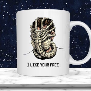 May include: White ceramic mug with a graphic of a facehugger from the Alien film series wrapped around a person's head. The mug has the text "I LIKE YOUR FACE" in black block letters.