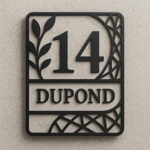 May include: Black metal house number sign displaying the number 14 and the name DUPOND. The rectangular sign features decorative cut-out designs, including a leaf branch and geometric patterns.