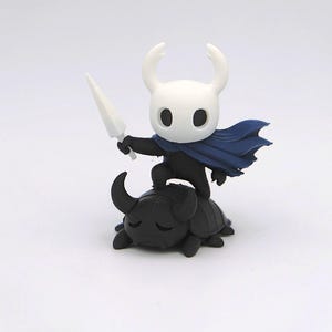May include: A figurine of a character from the video game Hollow Knight. The figure is white with black accents, a blue cape, and a white sword. It stands atop a black, beetle-like creature.