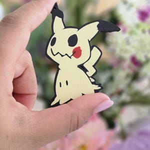Peut inclure: Une breloque plate d&eacute;coup&eacute;e en forme du personnage Mimikyu, pr&eacute;sentant un corps jaune p&acirc;le avec des contours noirs, des marques oculaires noires, une bouche dentel&eacute;e et un accent rouge circulaire sur la joue, avec un petit trou en haut.