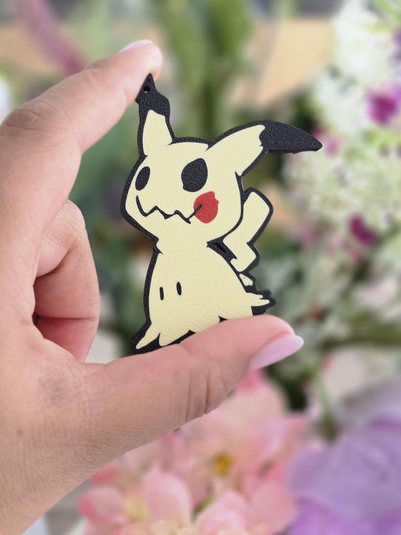 Peut inclure: Une breloque plate d&eacute;coup&eacute;e en forme du personnage Mimikyu, pr&eacute;sentant un corps jaune p&acirc;le avec des contours noirs, des marques oculaires noires, une bouche dentel&eacute;e et un accent rouge circulaire sur la joue, avec un petit trou en haut.
