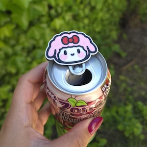 Op de afbeelding: Een roze en wit cartoonfiguur bliktopper met een rode strik is geplaatst op een frisdrankblikje. Het blikje is zilverkleurig en heeft een rood-wit etiket met groene bladeren. De achtergrond is wazig groen gebladerte.
