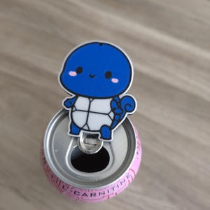Op de afbeelding: Een blauw-witte blikopener in de vorm van een schildpad met een stripfiguur met een rond hoofd, zwarte ogen en roze wangen. De opener is bevestigd aan de bovenkant van een roze drankblikje met de tekst CARNITINE duidelijk zichtbaar.