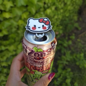 Puede incluir: Una lata de té helado Arizona con un adorno de Hello Kitty. La lata es roja y verde con las palabras "Arizona" y "Brooklyn Original". El adorno de Hello Kitty es blanco con un lazo y detalles rojos.