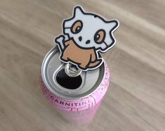 Peut inclure: Une canette de boisson rose en aluminium avec le mot CARNITINE imprimé sur le bord supérieur. Une breloque plate en forme de personnage de dessin animé, avec un masque de crâne blanc et un corps marron, est fixée à la languette argentée.