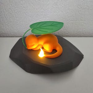 Può includere: Una statuetta di creatura arancione che riposa su una base di roccia grigio scuro. La creatura ha una coda curva con una fiamma gialla luminosa sulla punta ed è riparata da un grande baldacchino verde a forma di foglia che si inarca sul dorso.
