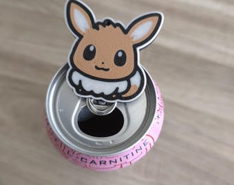 Peut inclure: Un charm représentant le personnage Eevee, de couleur marron et crème, fixé à la languette argentée d'une canette de boisson rose. Le texte L-CARNITINE est imprimé en noir sur le bord supérieur de la canette. Le charm a une texture visible.