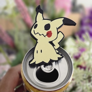 Peut inclure: Une figurine Mimikyu jaune p&acirc;le avec des accents noirs et une joue rouge, con&ccedil;ue pour s'adapter &agrave; la languette d'une canette de boisson en aluminium argent&eacute;. La silhouette est dentel&eacute;e et fantomatique avec des yeux sombres et une bouche cousue.