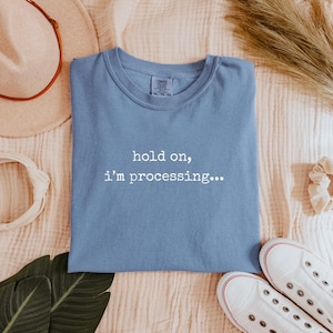 May include: A light blue t-shirt with the text "hold on, I'm processing..." in white. The shirt is folded and surrounded by a beige hat, white sneakers, and other accessories.
