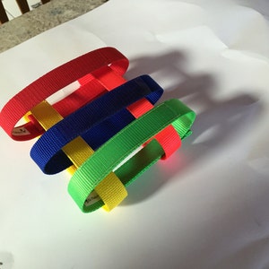 May include: Four woven colorful fabric bands in red, blue, green, and yellow. The bands are woven together to create a decorative pattern.